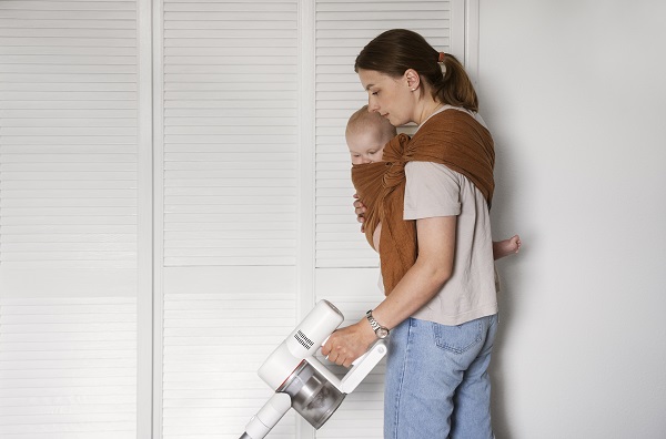 mama care își ține bebelușul într-un sling și care dă cu aspiratorul prin casă