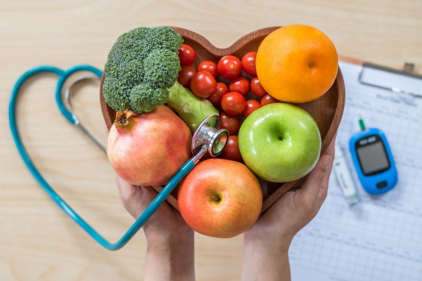persoană care ține în mână un castron în formă de inimă cu fructe și legume, alături de un stetoscop, promovare a ideii de alimentație care poate preveni diverse boli