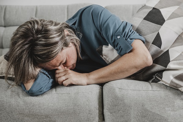 femeie matura imbracata cu bluza gri, care sta intinsa pe o canapea gri, care tine sub brat o perna cu nuante de gri si care se simte rau sau este stresata