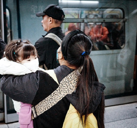 femeie cu rucsac in spate care tine in brate o fetita imbracata cu o vesta matlasata si care poarta o masca de protectie alba in timp ce se pregatesc sa se urce in metroul aglomerat