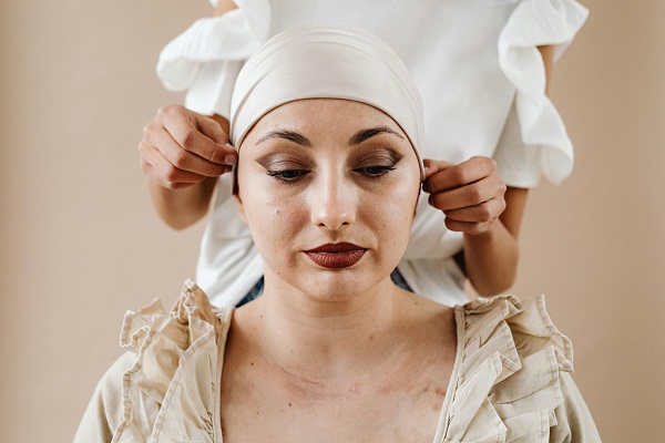 femeie trista, imbracata cu o bluza sau cu o rochie crem, cu vanatai sau cicatrici pe pielea din zona gâtului, privind in jos în timp ce o femeie care sta in spatele ei ii aranjeaza bandana alba