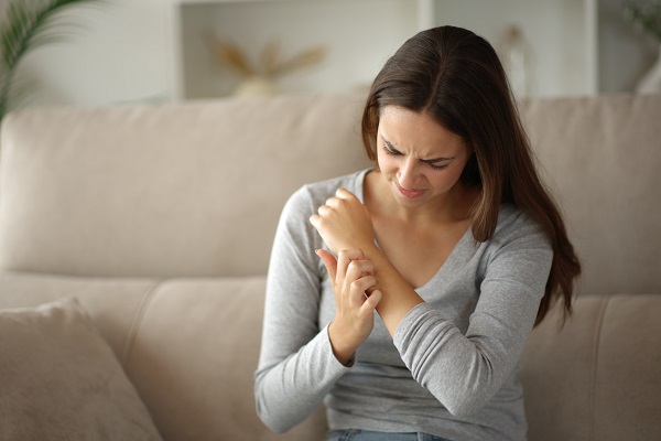 femeie tanara imbracata in bluza cu maneca lunga gri, stand pe o canapea crem, care resimte disconfort si se scarpina din cauza mancarimii de pe mana