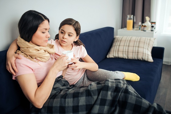 femeie imbracata in tricou roz care sta pe canapea si are la gat un fular bej in timp ce fetita ei sta langa ea, imbracata in tricou roz si colanti gri, si ii ofera o cana alba