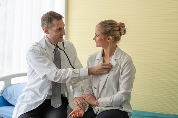 medic de varsta mijlocie zambitor, purtand halat alb, stand pe pat intr-un cabinet in timp ce masoara pulsul si o asculta cu stetoscopul pe pacienta lui,imbracata cu bluza alba cu maneca lunga, care se uita la el si ii zambeste   