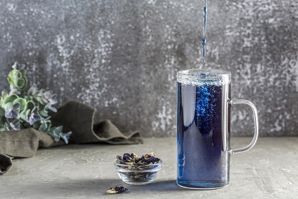 ceai albastru care se toarna intr-un pahar inalt de sticla, langa pahar aflandu-se un castronel de sticla cu flori albastre uscate de &bdquo;mazarea fluturilor&rdquo;, iar in spatele paharului o planta, o bucata de material textul inchisa la culoare si un perete gri