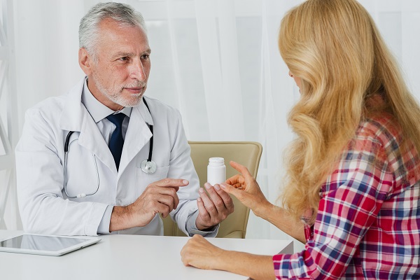 medic in varsta in halat, in cabinetul sau, in timp ce ii arata un flacon alb de medicamente pacientei sale, o femeie cu parul lung, blond, imbracata cu camasa in carouri rosii, albastre si albe