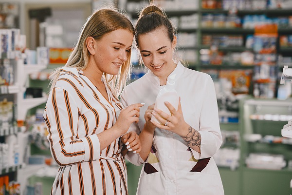 femeie tanara imbracata cu rochie alba cu dungi portocaliu cu negru zambitoare in timp ce se uita la un produs cosmetic intr-un recipient alba prezentat de o consiliera in frumusete zambitoare imbracata in uniforma alba cu cateva dungi in nuante de maro intr-un magazin cu produse cosmetice