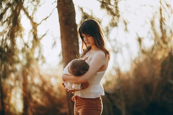 mama imbracata cu top alb cu bretele si pantaloni maro, care isi tine in brate bebelusul pe care il alapteaza intr-un cadru natural cu copaci scaldati in lumina soarelui
