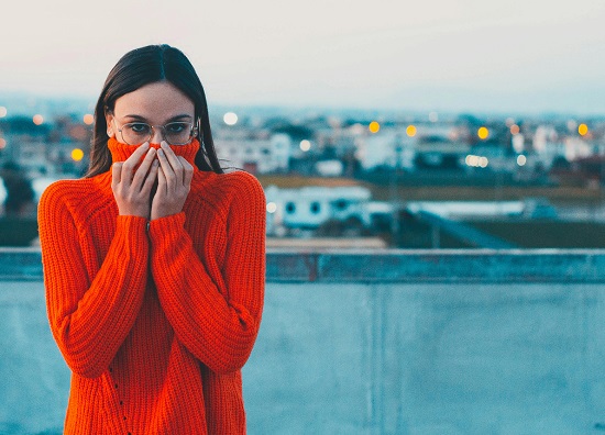 femeie cu parul lung, care poarta ochelari, ascunzandu-si chipul intr-un pulover rosu, in timp ce se afla in aer liber stand cu spatele la o terasa de pe care se vad in departare cladirile unei localitati si cateva lumini