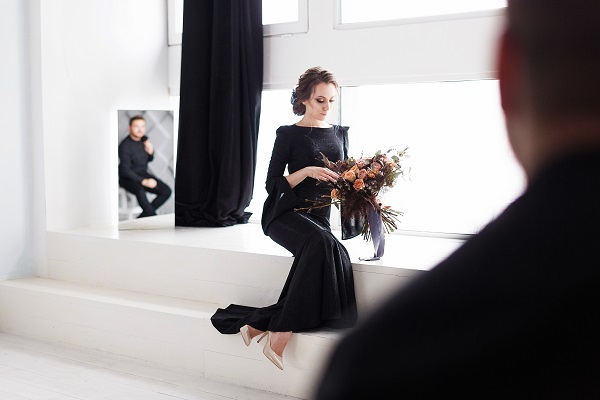 mireasa imbracata in rochie neagra,stand langa o fereastra cu privirea in jos,tinand in mana un buchet de trandafiri deschisi la culoare, in timp ce in oglinda din spatele ei se reflecta imaginea unui barbat imbracat in negru