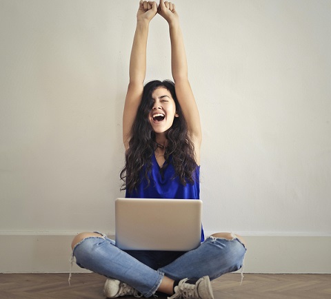 femeie tanara, cu parul lung, imbracata cu bluza albastra fara maneca si cu blugi rupti, entuziasmata, ridicandu-si bratele deasupra capului, in timp ce sta ghemuita, cu ochii inchisi, pe podea, tinand un laptop pe genunchi