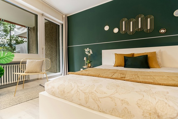 dormitor modern, avand peretele de langa pat vopsit cu verde inchis, cu decoruri aurii, patul fiind crem, cu perne decorative mustar si verzi, parchetul fiind crem, langa pat fiind o noptiera cu veioza si cu o floare alba, fereastra si un scaun bej cu pernuta bej