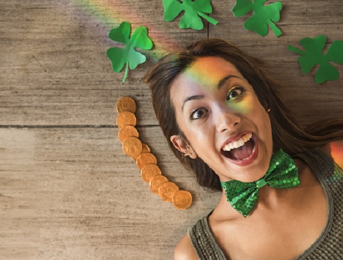 femeie tanara entuziasmata, stand intinsa pe o suprafata de lemn, imbracata intr-un maieu maro, avand la gat un papion verde cu paiete, avand langa ea mai multe monede si cativa trifoi cu patru foi din hartie