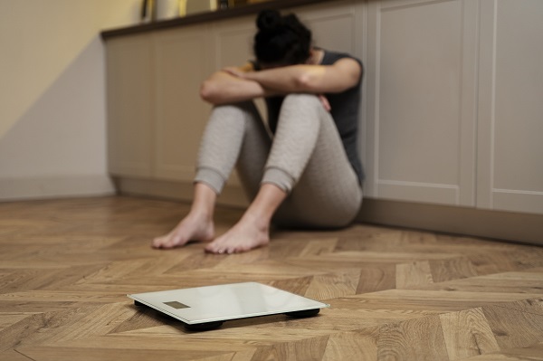 femeie cu parul strans, imbracata in pantaloni de trening gri deschis si bluza neagra fara maneca, tinandu-si capul intre maini si stand ghemuita pe podea in bucatarie, in timp ce in fata ei se afla un cantar