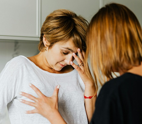 femeie cu parul scurt, imbracata in bluza alba, in timp ce isi duce mana la frunte intrucat nu se simte bine si se sprijina de un dulap alb din bucatarie, in timp ce alta femeie, imbracata in negru, vrea sa-i vina in ajutor 