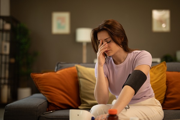 femeie tanara, imbracata in tricou lila si pantaloni crem, stand intr-o incapere a casei,pe o canapea maro cu pere decorative mustar si caramizii, simtandu-se rau, ducandu-si mana la frunte in timp ce isi masoara tensiunea arteriala cu un tensiometru pus pe o masuta