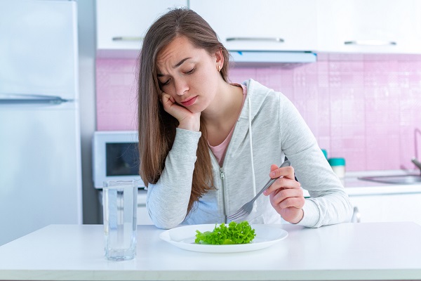femeie tanara, imbracata cu tricou roz si hanorac gri deschis, stand trista la masa, in fata unei farfurii cu o foaie de salata, in timp ce se afla in bucatarie