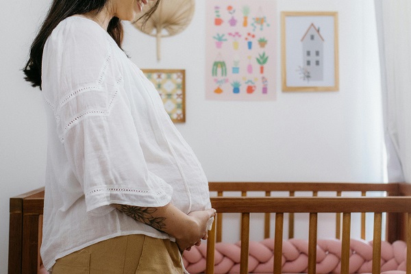 femeie insarcinata imbracata cu bluza alba si pantaloni mustar, zambind si atingandu-si burtica de gravida in timp ce sta langa un patut de copil cu roz intr-o camera de copil cu tablouri cu desene pe peretii albi