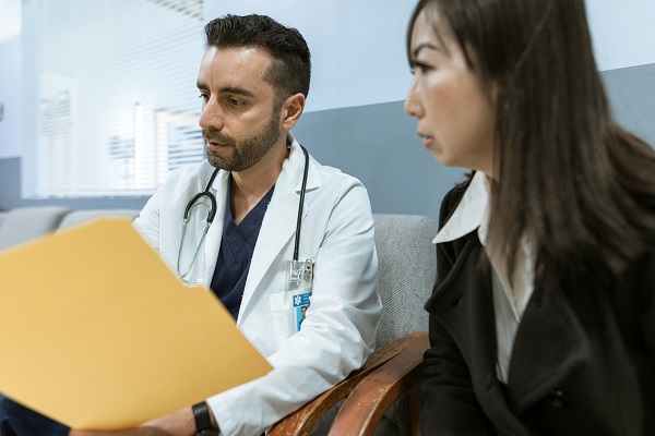 femeie imbracata cu bluza alba si haina de stofa neagra, stand pe o canapea langa un medic tanar imbracat cu uniforma bleumarin si halat alb, care examineaza un dosar medical pus intr-o mapa galbena