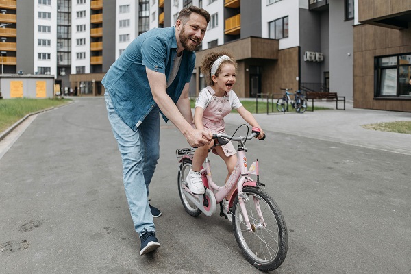 tata vesel, imbracat cu camasa de blugi si cu blugi, tinand ghidonul bicicletei fetitei sale, imbracata in roz, vesela, care incearca sa mearga cu bicicleta pe o strada printre blocuri