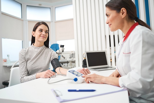 femeie tanara zambitoare, imbracata cu o bluza alba cu dungi negre, in timp ce o doctorita cu halat alb cu guler rosu ii masoara tensiunea arteriala la biroul ei, intr-un cabinet medical