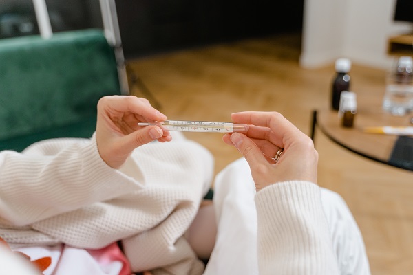 femeie imbracata in alb, invelita cu o patura alba, tinand in mana un termometru cu mercur in timp ce se afla acasa, langa ea fiind asezata o masuta cu medicamente