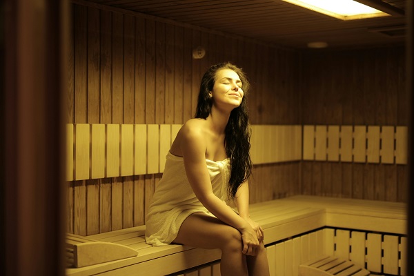 femeie tanara, zambitoare, avand pe ea un prosop alb, stand cu ochii inchisi si relaxandu-se la sauna