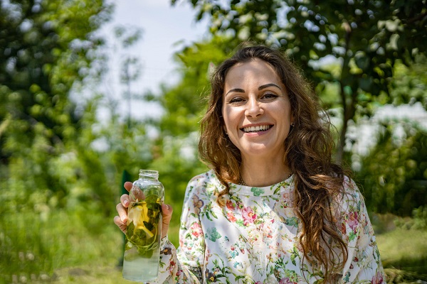femeie zambitoare, imbracata cu rochie sau cu bluza alba, cu imprimeu floral, care tine in mana o sticla cu apa infuzata, in timp ce se afla la umbra copacilor intr-o zi insorita