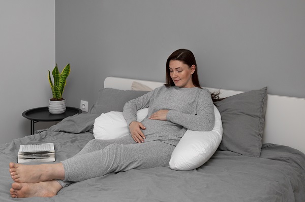 femeie insarcinata imbracata cu pijama gri, stand cu privirea in jos si mangaindu-si burtica de gravida in timp ce se sprijina pe o perna speciala pentru gravide alba, stand intr-un pat cu lenjerie gri, la picioarele ei aflandu-se o carte si langa pat o masuta rotunda cu o planta in ghiveci