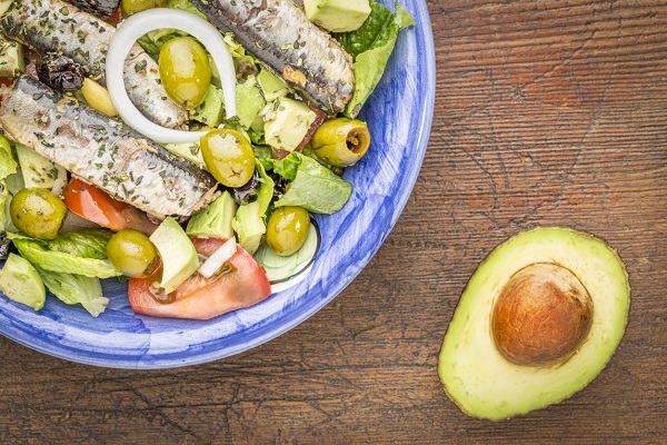 salata cu avocado, salata verde, masline verzi, sardine, rosii si ceapa pusa intr-un castron bleu, langa care se afla o jumatate de avocado