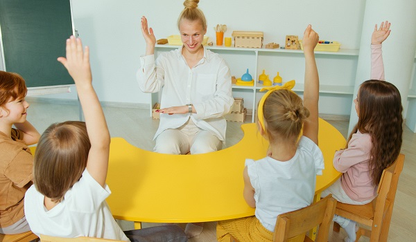 femeie tanara blonda, imbracata cu camasa alba si pantaloni crem, zambitoare, in timp ce sta pe un scaun cu spatele la o tabla si isi ridica bratele pentru a-si indemna si pe copiii din fata ei, care stau pe scaunele la o masa galbena, sa faca la fel, toti aflandu-se intr-o sala de clasa in timpul unei activitati