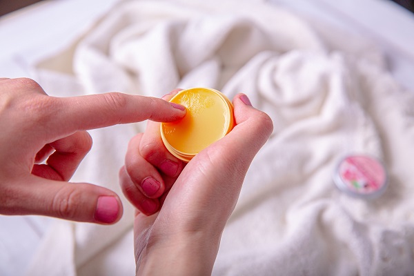 femeie cu manichiura cu oja roz, care incearca sa utilizeze un balsam de buze portocaliu, aflat intr-un recipient rotund tot portocaliu, pe fundal observandu-se un prosop alb pe care se afla alt recipient cu balsam de buze