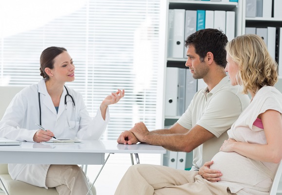 femeie insarcinata imbracata cu pantaloni si cu bluza crem, atingandu-si burtica de gravida in timp ce sta pe un scaun alaturi de sotul ei, imbracat cu tricou alb, in timp ce amandoi se afla intr-un cabinet medical, in fata unei doctorite in halat,cu stetoscop la gat, care gesticuleaza in timp ce le ofera exlicatii, langa biroul doctoritei aflandu-se o biblioteca cu dosare