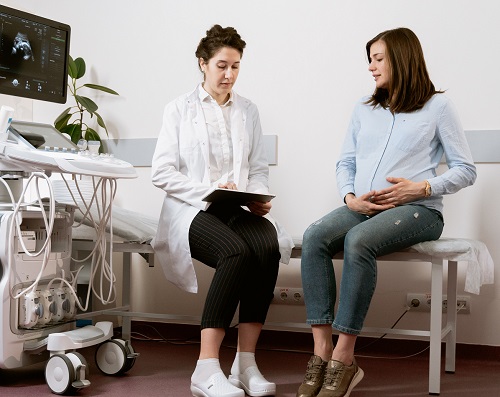 femeie tanara, insarcinata, imbracata cu camasa bleu si cu blugi, atingandu-si burtica de gravida,stand pe patul dintr-un cabinet de obstetrica-ginecologie, alaturi de o doctorita in halat alb si cu pantaloni negri, care tine in mana un material sau documente, oferindu-i explicatii tinerei insarcinate, langa ele aflandu-se echipamentul pentru ecografii