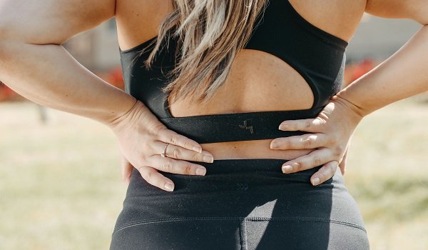 femeie imbracata in costum negru de sport, cu bustiera si colanti, tinandu-si mainile de o parte si de alta a spatelui, din pricina disconfortului sau durerii, in timp ce se afla in aer liber