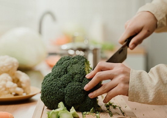 femeie purtand imbracaminte cu maneci lungi bej in timp ce incearca sa taie cu un cutit mare un broccoli pe un tocator de lemn in bucatatie, pe fundal sesizanduu-se un castron cu conopida, o varza si robinetul unei chiuvete
