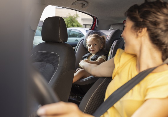 femeie tanara, imbracata cu tricou galben, avand centura de siguranta in timp ce se afla la volanul masinii, tinand de manuta si incercand sa distreze o fetita vesela, cu codite, care sta in scaunul special pentru copii in spate