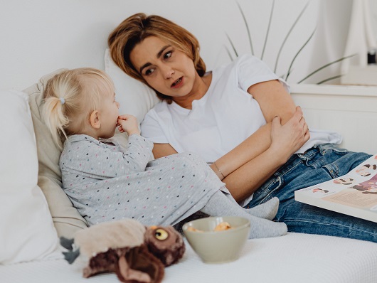 mama imbracata cu tricou alb si blugi, stand pe o canapea alba langa fetita ei blonda, cu codite, imbracata cu rochita gri cu buline, care rontaie ceva dintr-un castronel si are langa ea o jucarie de plus, in timp ce mama incearca sa ii dea explicatii in timp ce tine in poala o carte