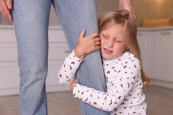 fetita blonda cu parul lung imbracata cu bluza alba cu floricele rosii, stand pe jos si plangand in timp ce tine cu mainile piciorul unei femei imbracate in blugi intr-un spatiu interior
