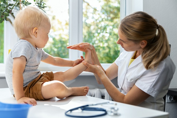 doctorita in uniforma alba cu maneca scurta, avand parul strans in coada, in timp ce examineaza talpa unui baietel blond, imbracat cu pantaloni scurt maro si cu tricou gri,care in sta in fundulet pe biroul ei alb din cabinet, birou aflat langa o fereastra
