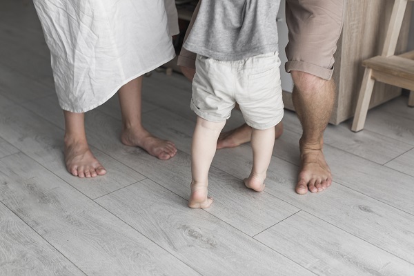 copil imbracat cu pantaloni scurti albi si cu tricou gri, care merge pe varfuri descult pe parchetul gri-bej de acasa, langa el aflandu-se o femeie desculta, in rochie alba si un barbat care il sustine, imbracat cu bermude bej, care isi indoaie genunchii si se sprijina, totodata, de mobila