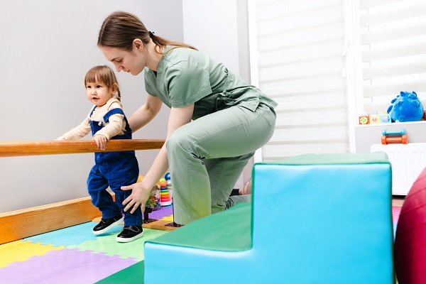 femeie kinetoterapeut, imbracata cu uniforma vernil, care se ocupa de o fetita cu codite, imbracata cu bluzita bej si cu salopeta albastra, care se sprijina de un perete alb si de o bara de lemn in timp ce face exercitii de mers intr-o sala special amenajata pentru kinetoterapie pentru copii