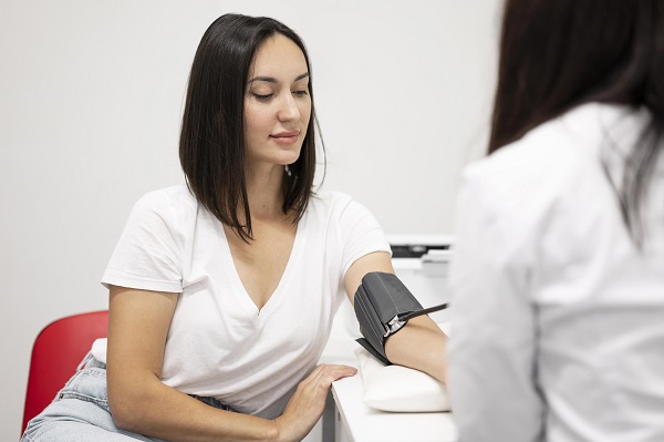femeie tanara imbracata cu tricou alb si blugi deschisi la culoare,, stand cu privirea in jos in timp ce sta pe un scaun rosu si i se masoara nivelul tensiunii arteriale cu un tensiometru de catre un cadru medical