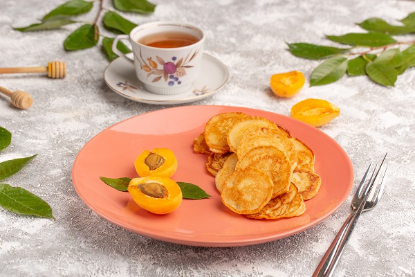 clatite pancakes asezate pe o farfurie intinsa roz alaturi de caise proaspete, langa farfurie aflandu-se o furculita, frunze, o cana alba cu ceai si jumatati de caise proaspete