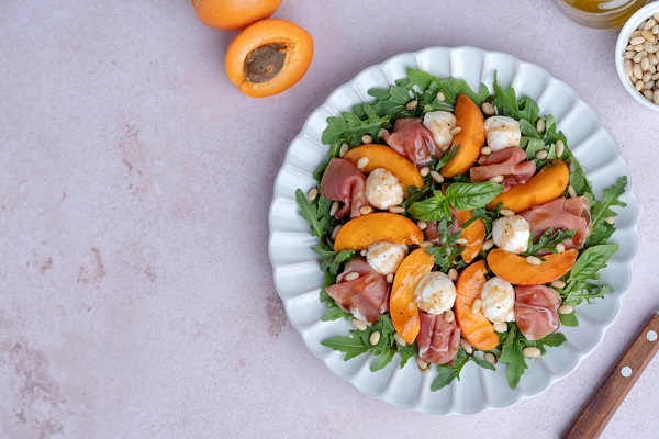 salata cu ruccola, seminte de pin, felii de caise, felii de prosciutto, busuioc, bile de mozzarella si cu otet balsamic pe o farfurie alba, langa care se afla un cutit si o parte dintre ingredientele folosite