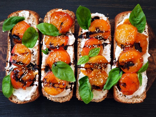 felii de paine unse cu crema cu branza, avand deasupra jumatati de caise caramelizate, frunze de busuioc, otet balsamic si susan, asezate pe un tocator de lemn,pe fundal negru