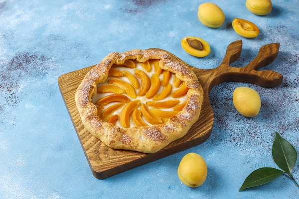 tarta rustica bine rumenita cu caise, pusa pe un tocator de lemn, langa care se afla caise proaspete si frunze de caise, pe un fundal albastru