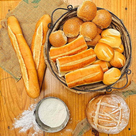 baghete puse pe un servet maroniu, alaturi de care se afla un cos metalic cu chifle si paini, o sita cu faina alba si o paine rotunda bine rumenita, cu faina deasupra, toate fiind asezate pe o suprafata de lemn