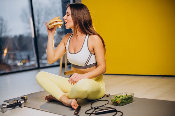 femeie tanara, imbracata cu bustiera alba cu gri sport si cu colanti galbeni, stand turceste pe un covoras maroniu, incercand sa muste dintr-un hamburger, in timp ce langa ea se afla greutati, o coarda, un recipient cu salata, femeia stand intr-o incapere cu pereti galbeni si cu ferestre mari