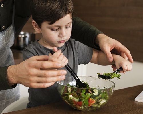 baietel imbracat cu tricou gri inchis, in timp ce un adult cu sort de bucatarie il ajuta sa amestece intr-o salata de legume intr-un castron de sticla pe o masa din bucatarie